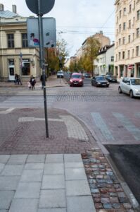 Šajā kadrā redzams jaunais (apakšā) un iepriekšējā gada (augšā) taktilā bruģa ieklāšanas veids. Iepriekš raupjais bruģis ieklāts ietves vidū, savukārt šogad ieklāts ietves malās.